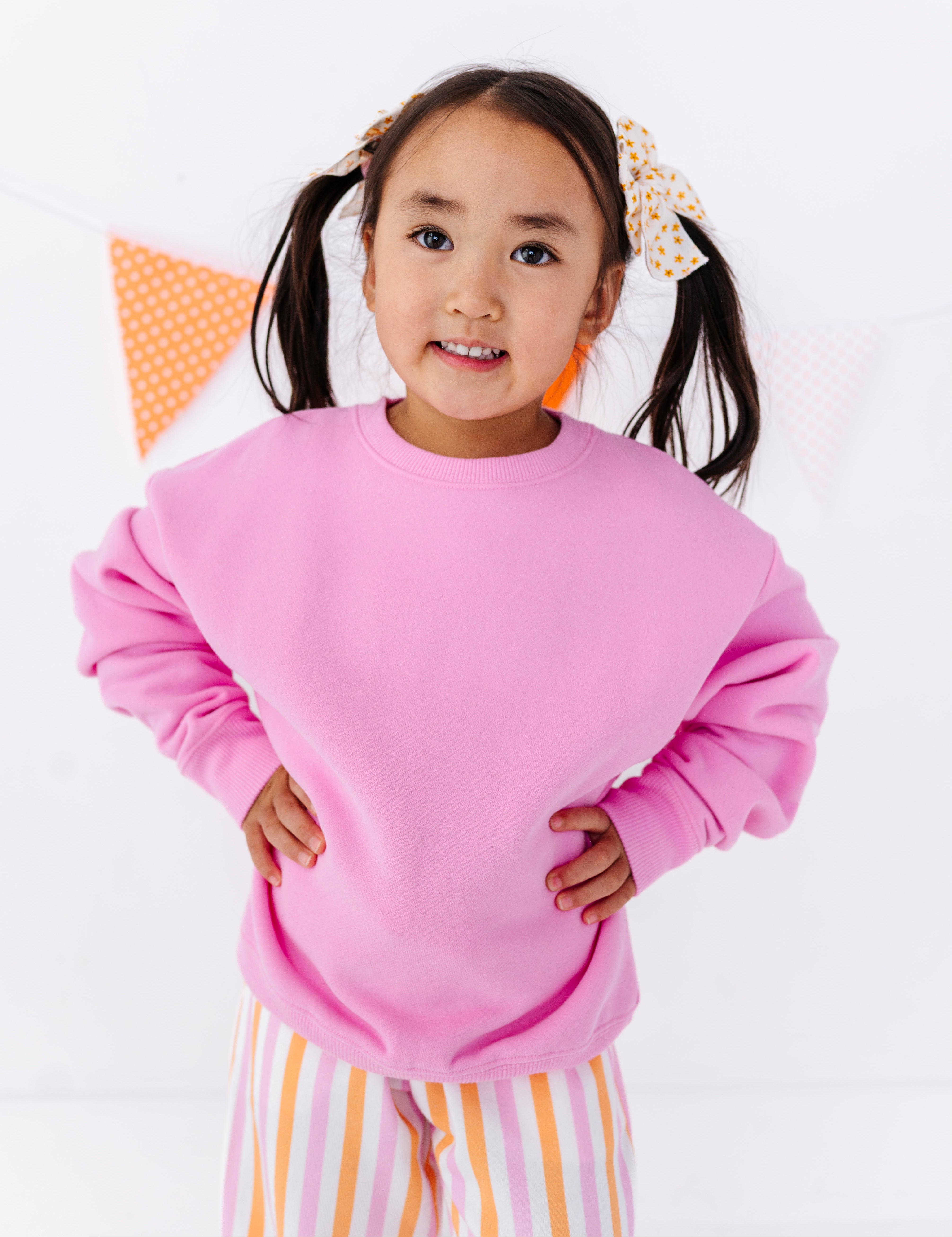 A young girl with dark hair in pigtails wearing an oversized pink sweatshirt and striped pants, posing with hands on hips against a white background with a subtle orange bunting decoration.
